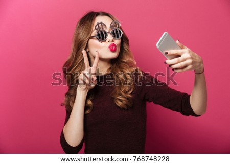 Close up portrait of a pretty funny girl wearing dollar shaped sunglasses taking a selfie isolated over pink background
