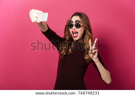 Close up portrait of a happy pretty girl wearing dollar shaped sunglasses taking a selfie isolated over pink background