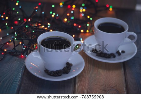 Festive still life with two cups of hot espresso. Coffee beans on saucers. Fiary Christmas lights in the back. Dark blurred bokeh. Dark wooden background. Close up.