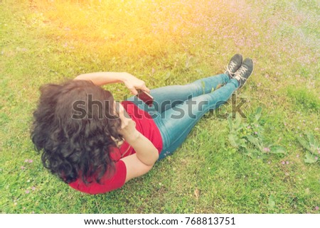 Brunette woman in a red T-shirt sitting on a field with flowers and taking pictures of her legs on the phone