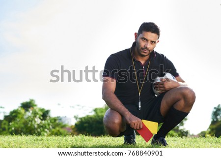 Full length portrait of confident mixed race soccer referee sitting on haunches with ball and penalty cards in hands and looking at camera, spacious football field on background