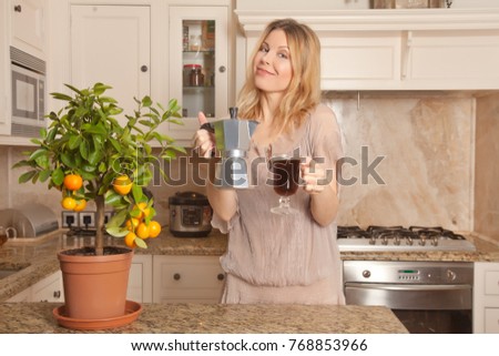 beautiful caucasian blonde housewife standing with a kettle and a cup of morning coffee in the kitchen at home