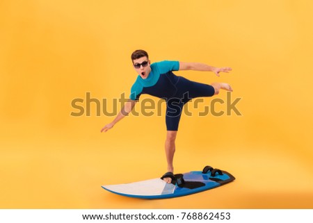 Picture of Funny surfer in wetsuit and sunglasses using surfboard like on wave and looking at the camera over yellow background