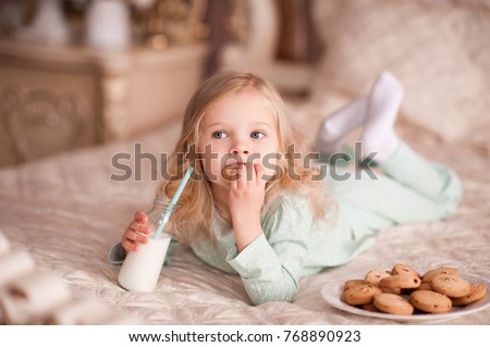 Funny girl 3-4 year old eating cookies and drinking milk in bed. Good morning. Breakfast.  Royalty-Free Stock Photo #768890923