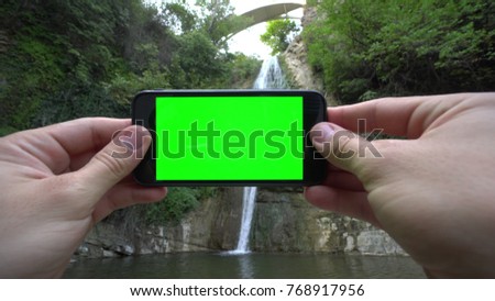 Hands holding (using) a smapthone with a green screen outside. Taking pictures of the waterfall. Close-up shot