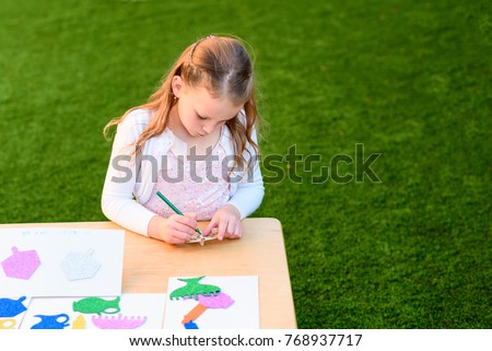A little girl paints a wooden craft and pastes stickers outdoor. Child doing handicrafts in the park at sunset.On the paper traditional Chanukah symbols - wooden dreidels , menorah, candles, oil jar.