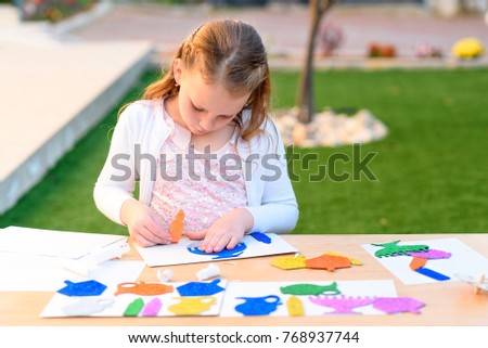 Little girl create a greeting card image of the Jewish holiday of Hanukkah. Kid pastes stickers - menorah, candles, dreidel, oil jar on paper on wooden table. Needlework, crafts for children.