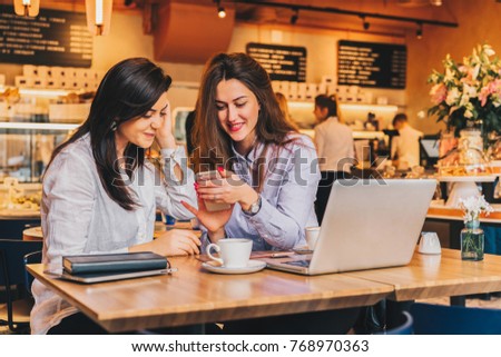 Two young happy women are sitting in cafe at table in front of laptop, using smartphone and laughing. On table paper notebook and cup of coffee. Girls are blogging, working, studying, learning online.