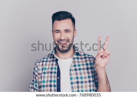 Portrait of young, smiling man in checkered shirt with stubble, bristle gesturing v-sign over grey background