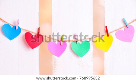 Cheerful image with colorful paper hearts with place for text on them, tied to a linen string with wooden clips, with a white wooden fence in the background.