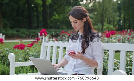 Beautiful brunnette lady sitting on a white bench in the park and using a laptop and a telephone. Medium shot