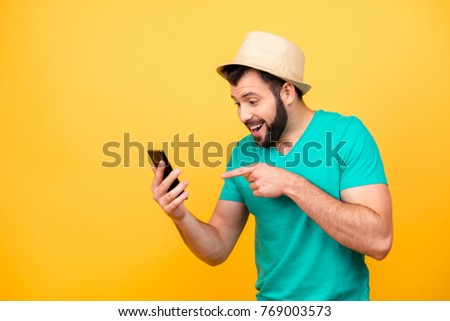 Ha-ha! So funny! Portrait of happy excited crazy man with stubble wearing hat and green tshirt, he is pointing on his smartphone and rejoicing