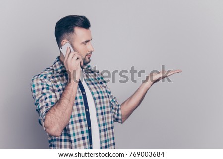 Side view portrait of  attractive, half-turned man with bristle in checkered shirt speaking on smart phone, gesturing, looking at copy space on palm, presenting new product, over grey background