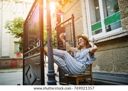 Portrait of a gorgeous model in striped overall sitting on the chair outdoor and taking selfie.