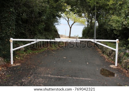 Strong white metal ramp locked with two padlocks on each side completely blocking entrance through paved road to local beach with green trees, traditional stone wall and sea in background