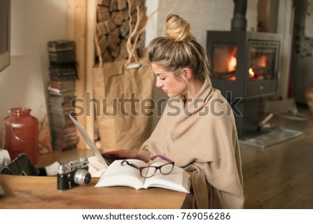 Young beautiful woman in the living room sits on the floor and communicates with friends over the computer, covered with a blanket for the cold in the back of the fireplace