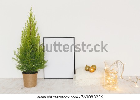 Composition of black frame with place for text, little Christmas tree in a pot, fur plaid, vase with Christmas garland lights and Christmas balls and on woden table. Mock up.
