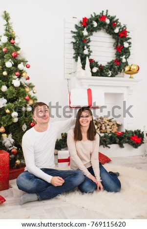Happy funny young cheerful couple in casual clothes sitting in light room at home with decorated New Year tree and gift box on head. Christmas good mood. Family, love and holiday 2018 concept