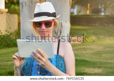 Young student woman sitting on a green grass in a park and holding a tablet computer outdoor leaning on a palm tree trunk 