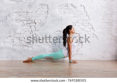 Side view - beautiful young brunette woman in gym clothes makes exercises for stretching  back and legs in an empty gym on white brick wall background. Space for text Royalty-Free Stock Photo #769188505