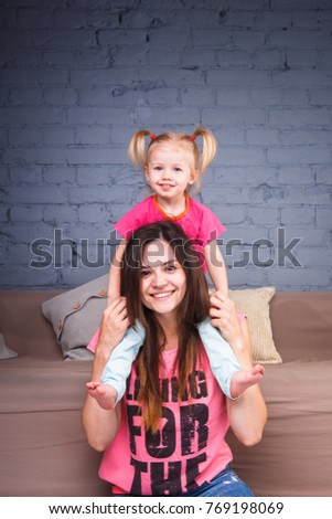 a young slender beautiful mother with a blond daughter on her shoulders play, am happy and indulge in the committees near the sofa on the wooden floor. Family values