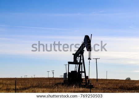 Silhouette of oil-gas pump jack out in field behind barbed wire fence silhouetted against blue sky with whispy clouds - with electric poles