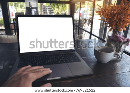 Mockup image of a hand using and touching laptop with blank white screen , coffee cup and flower vase on wooden table in cafe