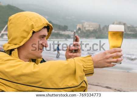 Adult man in a yellow jacket takes a photo of beer in a glass on the phone