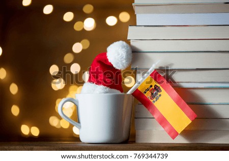 White cup and Christmas hat with Spain flag near books on fairy lights background