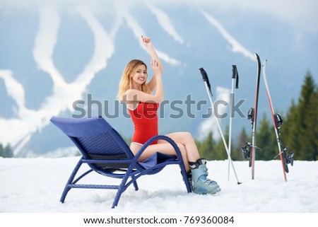 Smiling woman skier wearing red bodice, posing on a blue deck chair near skis and poles at ski resort. Mountains and ski slopes on the background. Ski season and winter sports concept