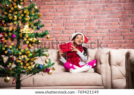 Cute Little Indian/Asian Girl Celebrating Christmas while wearing Santa Clause dress and Hat, sitting over Sofa with gifts and Tree in the background against red brick wall, dreamy lighting