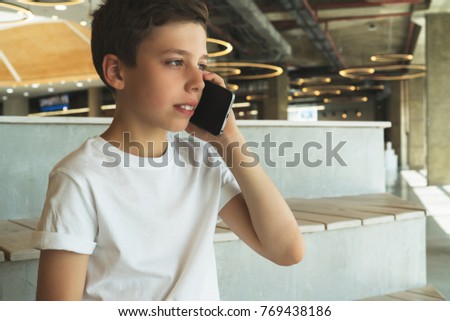 Boy in white t-shirt is sitting indoors and talking on his mobile phone. A teenager uses a cell phone, calling, phoning.