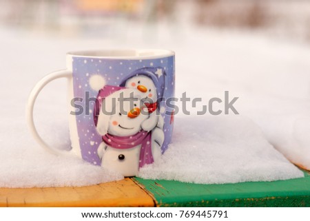 Tea in the winter in the snow. A picture of a snowman on a mug. Two funny snowmen are depicted on a mug of tea. A cup of tea in winter for the new year. A mug of tea with snowmen and snow.
