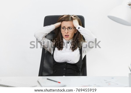 The beautiful tired perplexed and stress brown-hair business woman in suit and glasses sitting at the desk, working at contemporary computer with documents in light office on white background