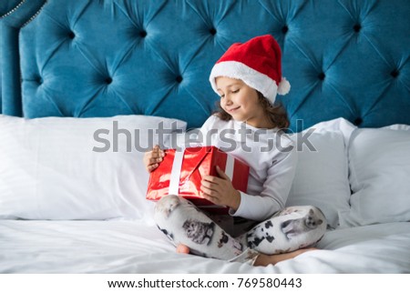 Happy excited little girl in Santa's hat holding gift box, looking at camera while sitting on bed