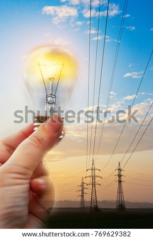electricity expenses and costs savings - electrical energy concept - hand holding a bulb against power station with pylons at sunset