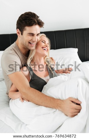Happy blonde lady laughing while lying in bed with her cheerful brunette man and hugging
