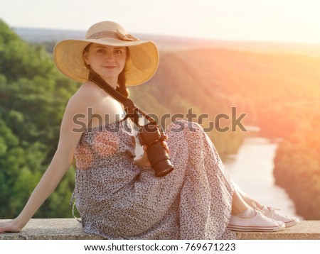 Girl in hat sits on hill with camera on the background of forest and winding river