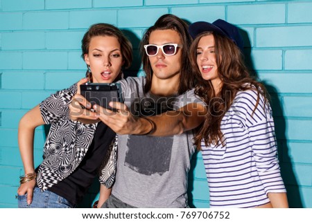 Best friend hipsters wearing stylish bright outfits and having great time. Handsome boy with two beautiful ladies taking selfie self portrait pictures with film camera in front of blue brick.