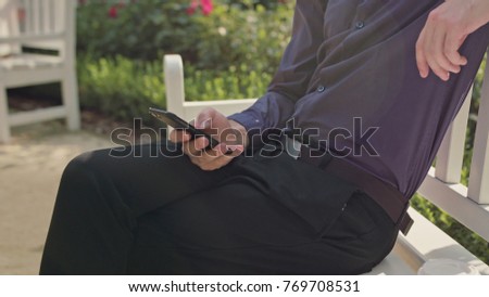 A young man sitting on a white bench in the park and using a phone. Close-up shot