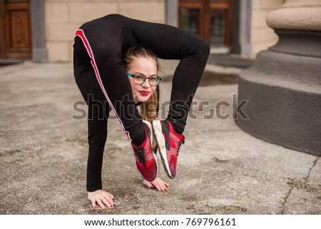 A young, slender girl in tight black tracksuit, doing complex acrobatics in the street in cloudy weather. Very good stretching and flexible back.