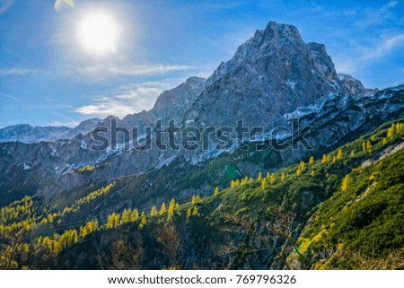 An autumn scenery in the austrian alps with two very high mountains partly covered with snow, steep rock walls and grassland with little trees, green and yellow, pictured against the sun light