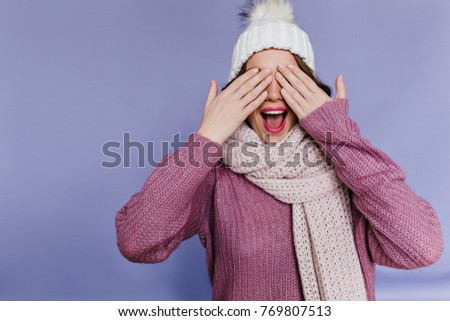 Spectacular woman in trendy knitted scarf covering eyes with hands. Indoor photo of laughing winsome woman in hat and sweater funny posing in front of purple wall.