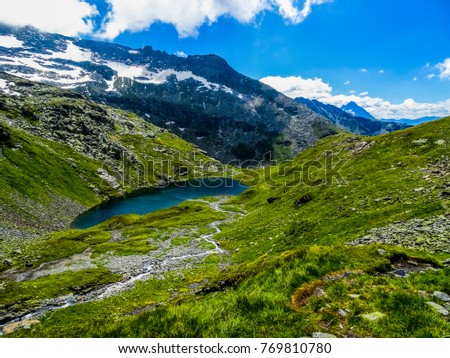 Green grass mountain sides surrounding a dark turquoise coloured lake in the middle of the austrian alps, with a high mountain side covered with snow and ice, all below a blue sky