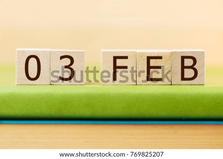 Wooden cube shape calendar for FEB 3 on green book, table. 