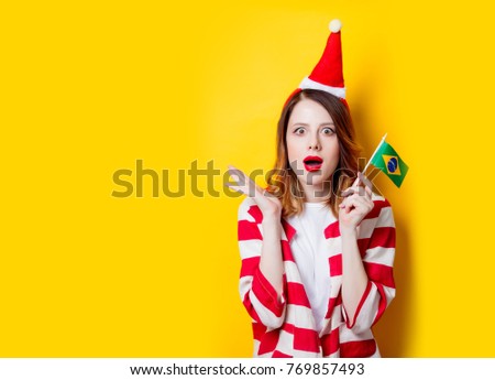 Portrait of young redhead woman in Santa Claus hat and striped shirt with Brasil flag on yellow background. Christmas time