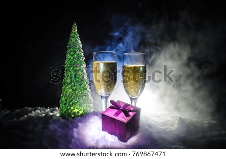 New Years Eve celebration background with pair of flutes and bottle of champagne with christmas tree on snow on dark background. Selective focus