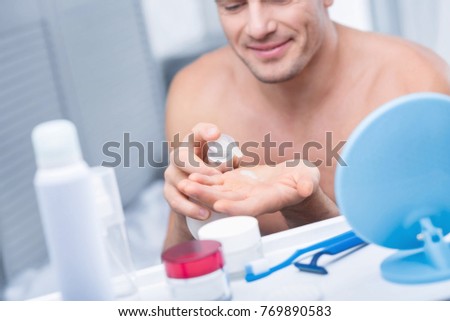 Exact dosage. Close up of happy energetic man pressing bottle for getting cream while looking down and smiling