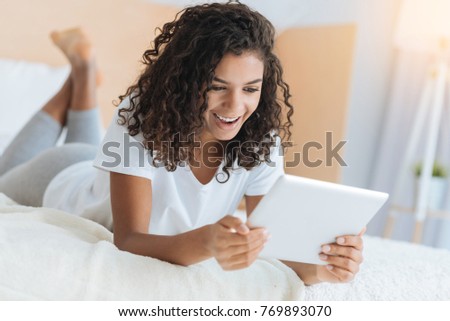 Excited time. Radiant young lady lying on a bed and grinning broadly while looking at a screen of her tablet computer at home.