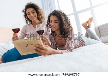 Recalling memories. Low angle shot of two joyful ladies grinning broadly while holding their glasses of wine and focusing their attention on a framed photo at home.